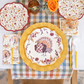 Elevate your festive harvest meal or gathering with the Amber Harvest Table Setting by Sophistiplate. This colorful autumn tablescape features turkey-themed designer disposable plates, a yellow charger, floral napkins, and plastic flatware, all beautifully arranged alongside an orange flower centerpiece on a plaid runner and crisp white tablecloth.