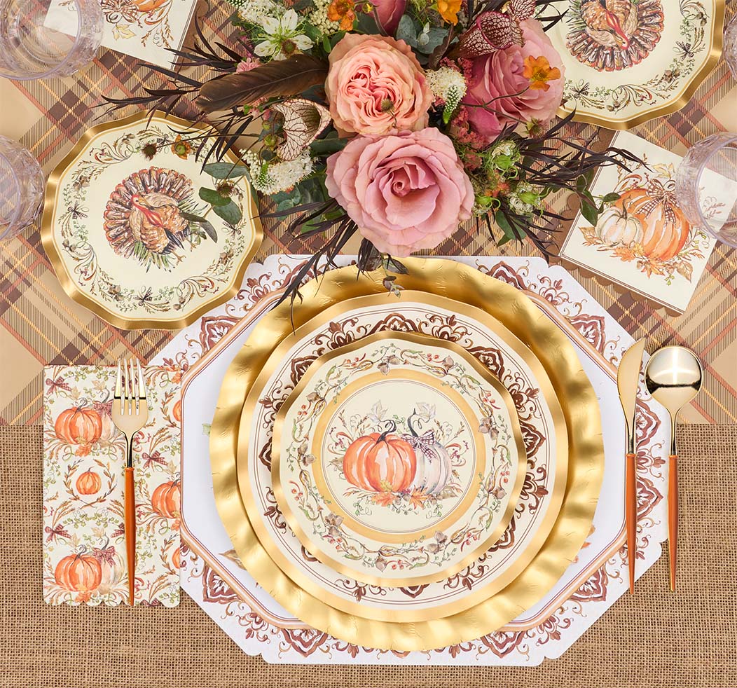 A fall-themed table setting features the Turkey & Gourds Table Setting Bundle by Sophistiplate LLC, including designer disposable plates adorned with turkey and gourd motifs, gold cutlery, plaid and burlap placemats, and a floral centerpiece with pink roses and autumnal blooms. The distinctive turkey design appears on both the top plate and matching napkin.