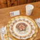 A Sophistiplate LLC Turkey & Gourds: Thanksgiving Table for 16 place setting featuring a decorative turkey heavy duty paper plate on a gold charger, a striped paper cup, a place card with a bow, and a napkin decorated with gourds.