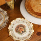 A decorative Turkey & Gourds: Thanksgiving Table for 16 setting featuring a turkey illustration on a disposable party plate, placed on a wooden surface next to a whole pie and a glass filled with gold cutlery.
