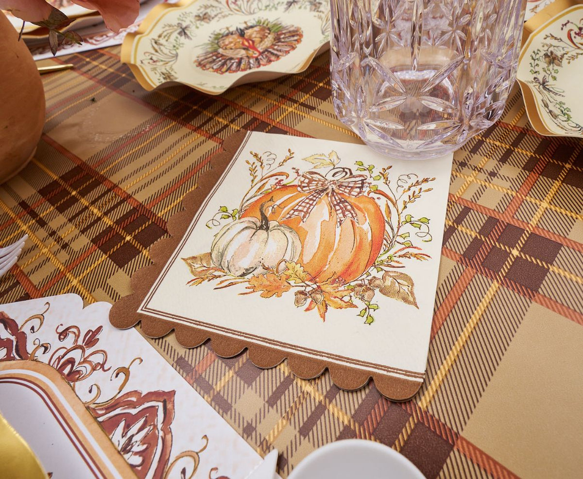 The Turkey & Gourds Table Setting Bundle by Sophistiplate LLC features festive napkins adorned with pumpkin illustrations and a bow, arranged on a plaid tablecloth alongside stylish disposable plates, a clear glass, and autumn-inspired tableware, creating a cozy Thanksgiving or fall-themed atmosphere.