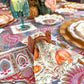 A Turkey & Gourds Guest Towels/20pk harvest pattern napkin decorated with pumpkins and autumn leaves, tucked into a turkey-shaped napkin ring on a patterned tablecloth.