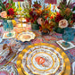 Turkey & Gourds: Thanksgiving Table for 16 set on a patterned tablecloth with gold chargers, teal glasses, and floral centerpieces with candles, featuring paper plates decorated with pumpkin illustrations.