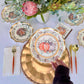 A hand sets the Sophistiplate Turkey & Gourds Table Setting, featuring a designer paper plate, at a fall-themed table with matching napkin, cutlery, and a floral and succulent centerpiece.