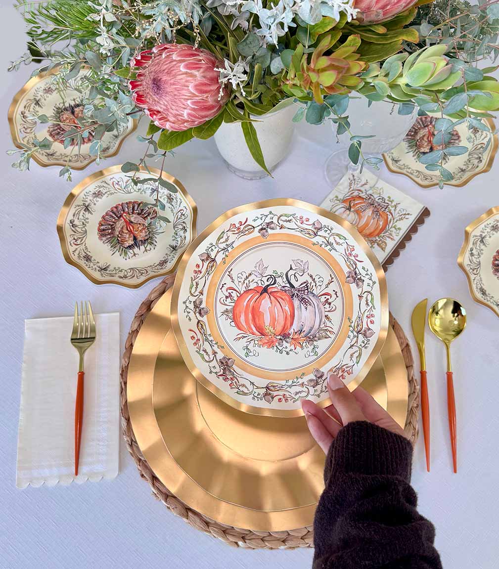 A hand sets the Sophistiplate Turkey & Gourds Table Setting, featuring a designer paper plate, at a fall-themed table with matching napkin, cutlery, and a floral and succulent centerpiece.