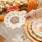 A hand displays the Sophistiplate Turkey & Gourds Table Setting paper plate featuring a turkey design above a festive Thanksgiving table with matching autumn plates, pumpkins, candles, and a patterned napkin.