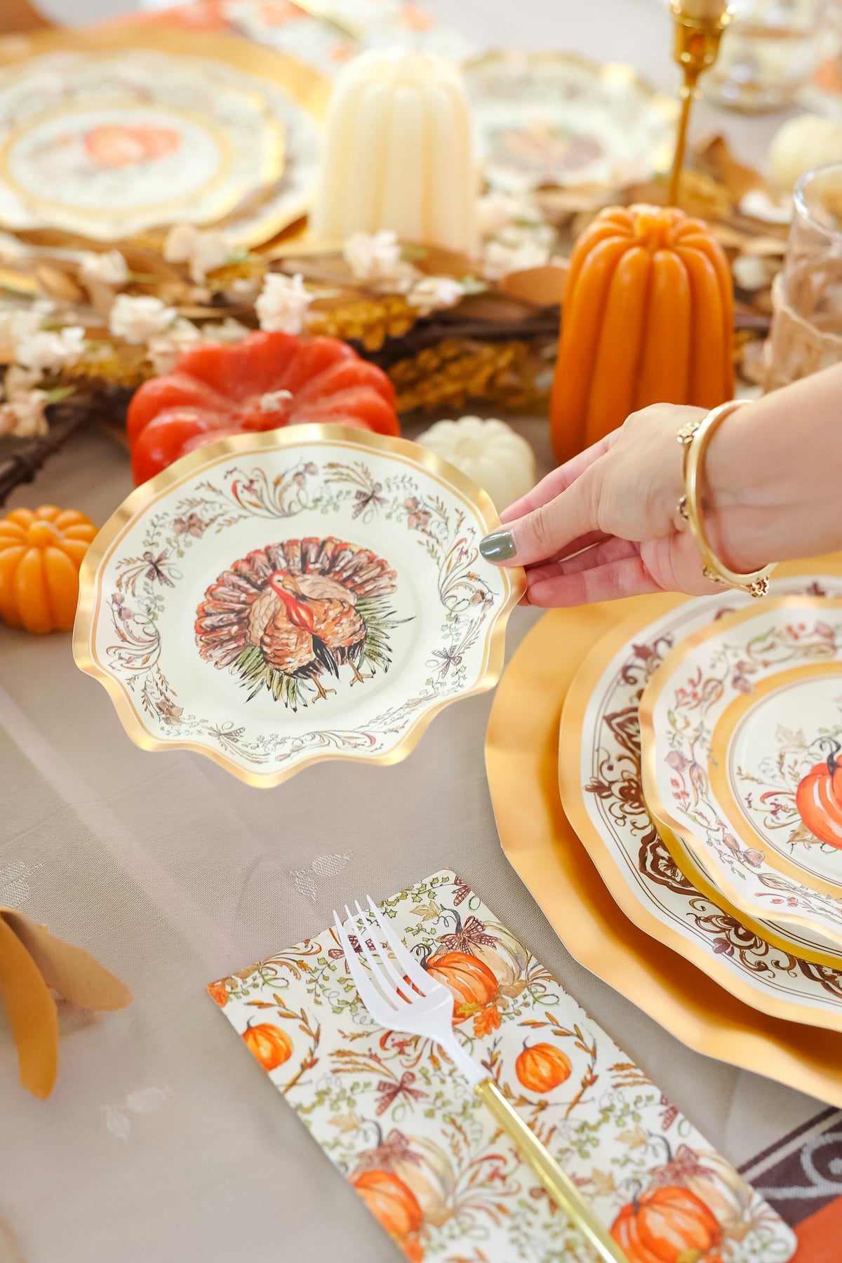 A hand displays the Sophistiplate Turkey & Gourds Table Setting paper plate featuring a turkey design above a festive Thanksgiving table with matching autumn plates, pumpkins, candles, and a patterned napkin.